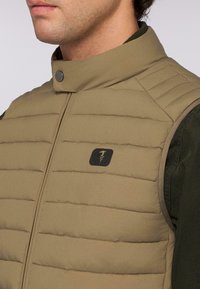 Hnědá izolační vesta s horizontálním prošívaným vzorem a stojáčkovým límcem. Má logo na hrudi a hladkou texturu materiálu.