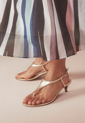 Füße, die goldene, niedrigabsätzige Sandalen mit Riemchen unter einem fließenden Rock mit vertikalen Streifen in Schwarz, Weiß, Blau und Pink tragen.