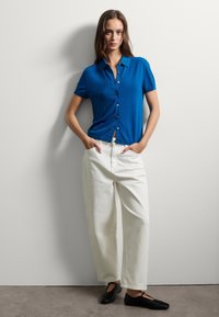 Camicia a maniche corte con bottoni di un vivace blu. Tessuto liscio. Abbinata a pantaloni bianchi larghi e scarpe nere basse.