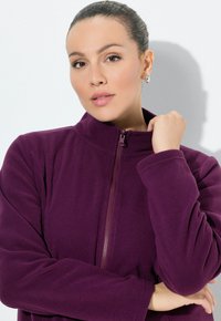 Donna con i capelli raccolti, che indossa orecchini a cerchio d'argento e una giacca in pile viola con zip, in posa contro un muro bianco.