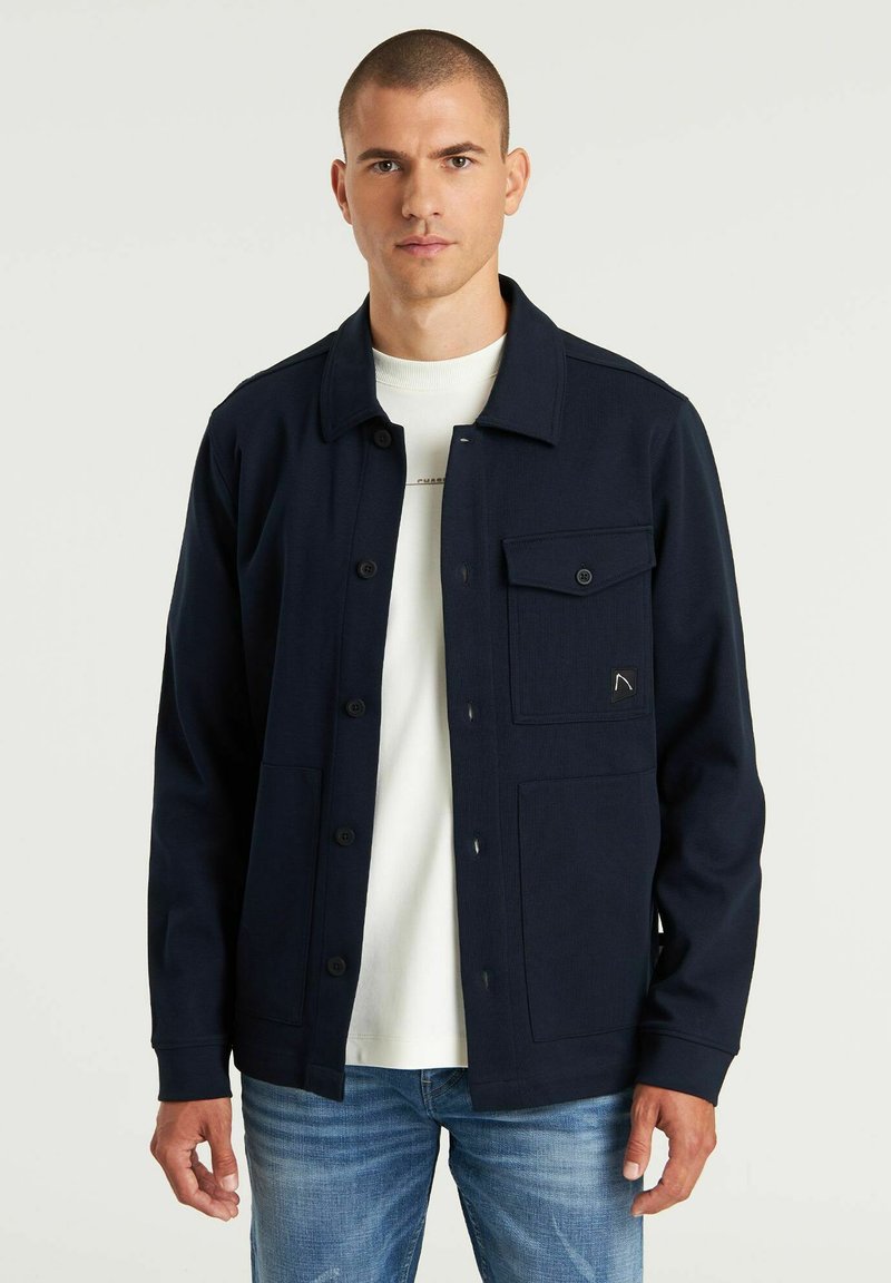 Marineblaues Hemd mit Knöpfen, ausgestattet mit vier vorderen Taschen, aus einem strukturierten Material gefertigt. Kombiniert mit einem weißen Rundhals-T-Shirt und blauen Jeans.