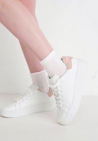 Sneakers bianchi con una texture liscia, caratterizzati da un accento rosa pallido sul tallone. Design con lacci e suola spessa, indossati con calze bianche.