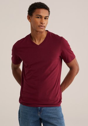 Ung mand med kort krøllet hår iført en bordeaux V-hals T-shirt og blå jeans mod en ensfarvet beige baggrund.