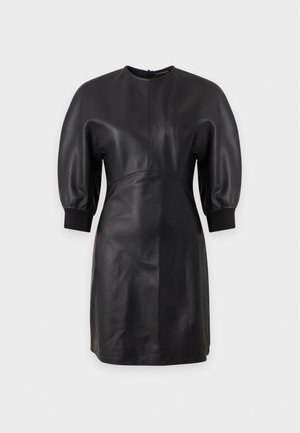Femme portant une courte robe en cuir noire avec des manches trois-quarts bouffantes et des chaussures à talons hauts noires, debout devant un fond clair uni.