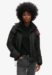 Sort dunjakke med høj krave, lynlås og flere lommer. Har et logo-patch på skulderen. Bæres over en sort sweater.