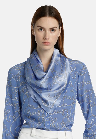 Blauwe zijden sjaal met een satijnachtige afwerking, met een ingewikkeld touwpatroon in zilver, gedragen gedrapeerd om de nek over een gestippeld shirt.