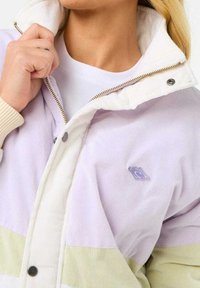 Giacca di velluto a coste in lavanda e bianco con dettagli verdi; presenta una chiusura con zip, bottoni a pressione e un piccolo patch con logo sul petto.