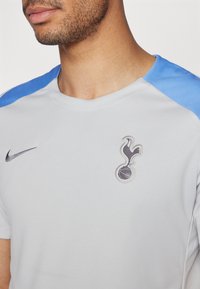 Sivý športový dres s modrými akcentmi na pleciach. Obsahuje logo Nike a vyšitý tímový emblém na hrudi. Hladký, syntetický materiál.