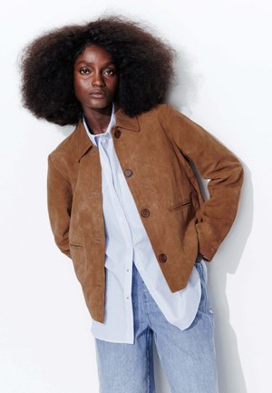 Veste en daim brun avec fermeture à bouton, deux poches avant ; superposée à une chemise à rayures bleues et blanches et un jean délavé.