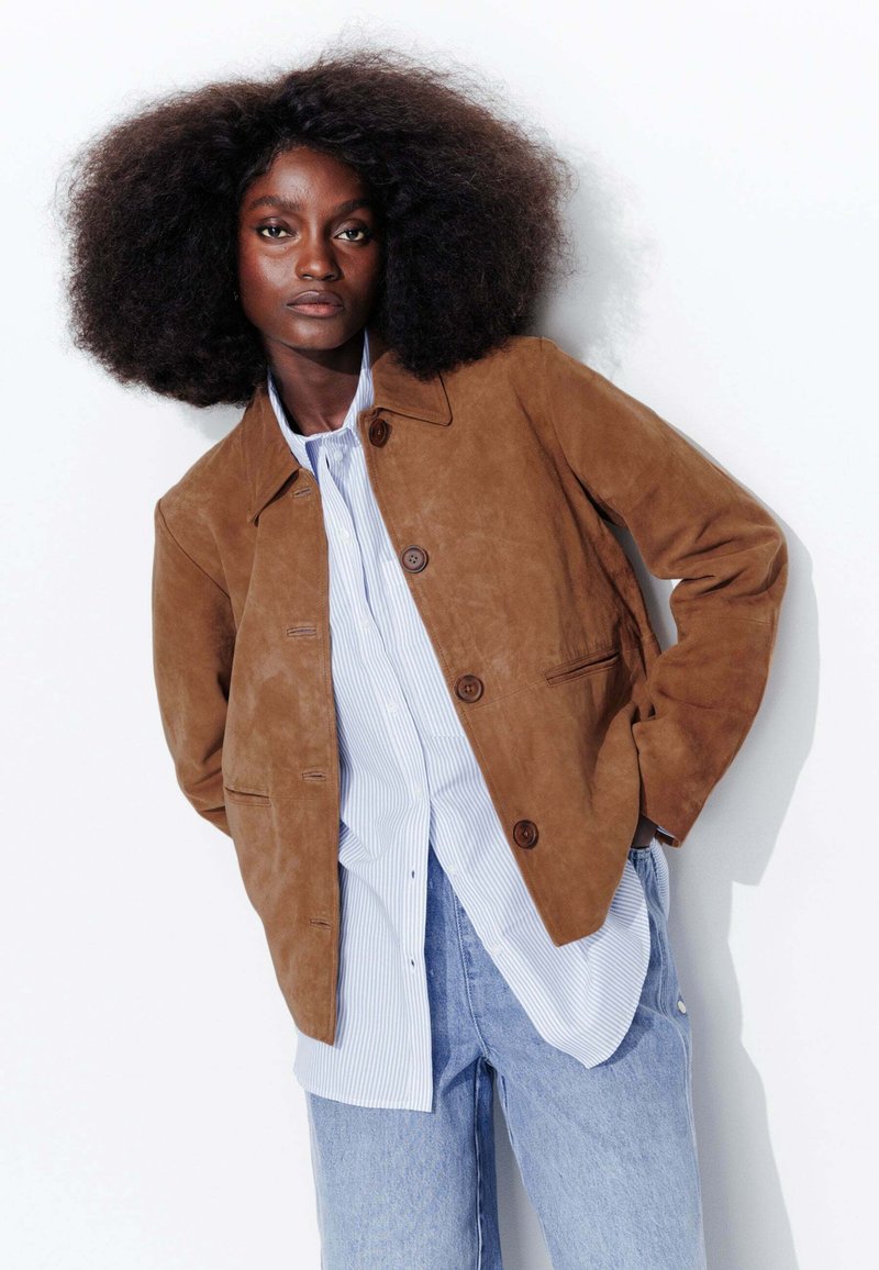 Veste en daim brun avec fermeture à bouton, deux poches avant ; superposée à une chemise à rayures bleues et blanches et un jean délavé.