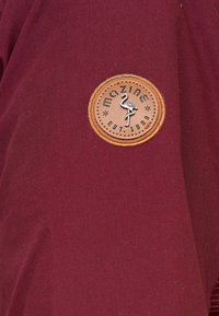 Patch circulaire marron avec un flamant rose et le texte "mazine EST. 1999" cousu sur un tissu rouge foncé.