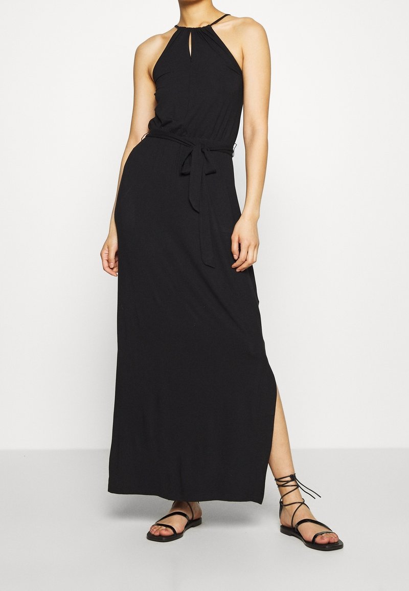 Femme portant une robe maxi noire sans manches avec un col halter, une ceinture à la taille, une fente latérale et des sandales plates noires à lacets.