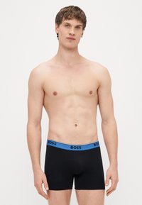Μαύρα ανδρικά εσώρουχα βρακάκι με μπλε λωρίδα που φέρει το λογότυπο «BOSS» στη μέση. Το ύφασμα φαίνεται απαλό και ελαστικό, εφαρμόζοντας στενά στο σώμα.