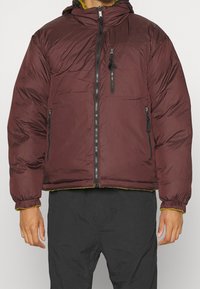 Burgundy vadderad jacka med huva, dragkedja, elastiska ärmslut och gul kant. Har en bröstficka och två sidofickor.