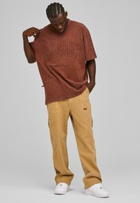 Brun oversized t-skjorte med hevet tekst, paret med beige cargo-bukser med sidelommer og detaljert logo. Hvit sneakers fullfører antrekket.