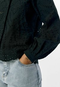 Zwarte geborduurde blouse met ingewikkelde uitsnijdingen en een met knopen sluitende mouw. Combinatie met lichtblauwe jeans, die textuur en pasvorm tonen.