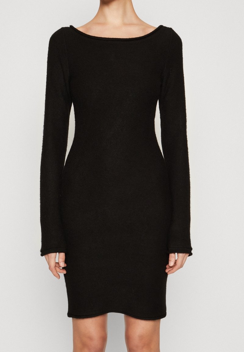 Femme portant une robe noire cintrée à manches longues avec un large décolleté, debout devant un fond clair uni.