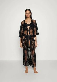 Ženska stoji bosa, oblečena v prosojni črni cvetlični čipkast dolg kimono preko črnega spodnjega perila, na enobarvni podlagi.