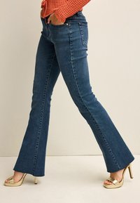 Dunkelblaue, ausgestellte Denim-Jeans mit glatter Textur, ausgestattet mit Fronttaschen und Nähdetails. Kombiniert mit goldenen High Heels.
