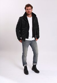 Černá kožená bomber bunda s kapucí, bílý tričko, světle modré džíny a černé kotníkové boty. Model pózuje na jednobarevném pozadí.