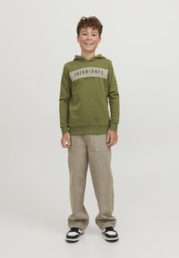 Sudadera verde con capucha y diseño de rayas blancas con texto, combinada con pantalones cargo beige y zapatillas negras y blancas. Un look simple y casual.