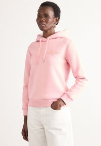 Rožinė hoodie pagaminta iš minkštos medžiagos, su priekiu esančiu logotipu ir su raišteliais reguliuojamu kapisonu. Modelis dėvi kreminės spalvos kelnes, kurios demonstruoja glotnias tekstūras.