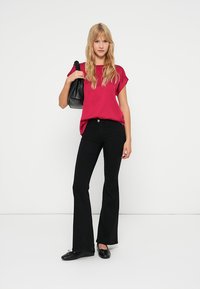 Tettsittende magenta topp med rullede ermer, kombinert med svarte flagrende bukser. Modellen holder en teksturert svart veske og har på seg svarte leiligheter.