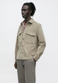 Beige corduroy overhemd met twee borstzakken, een kraag met knopen en lange mouwen, gecombineerd met grijze broeken. Gladde textuur, losse pasvorm.