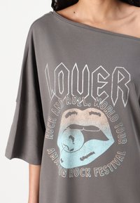 Pilkas per didelis T-shirt su apvaliu kaklu, su grafikiniu dizainu, vaizduojančiu lūpas ir tekstą balta ir pastelinių spalvų. Minkšta audinio tekstūra.