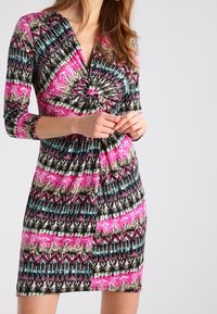 Kurzes Kleid mit tiefem V-Ausschnitt, mehrfarbigen geometrischen und abstrakten Mustern aus überwiegend pinken, schwarzen und türkisfarbenen Stoffen.