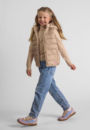 Ragazza sorridente che cammina, indossando un gilet trapuntato beige, un maglione beige, jeans blu e sneakers viola pastello su uno sfondo chiaro e semplice.