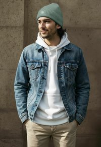 Denim jacket in blue with two chest pockets, layered over a white hoodie. A ribbed, light green beanie completes the outfit.