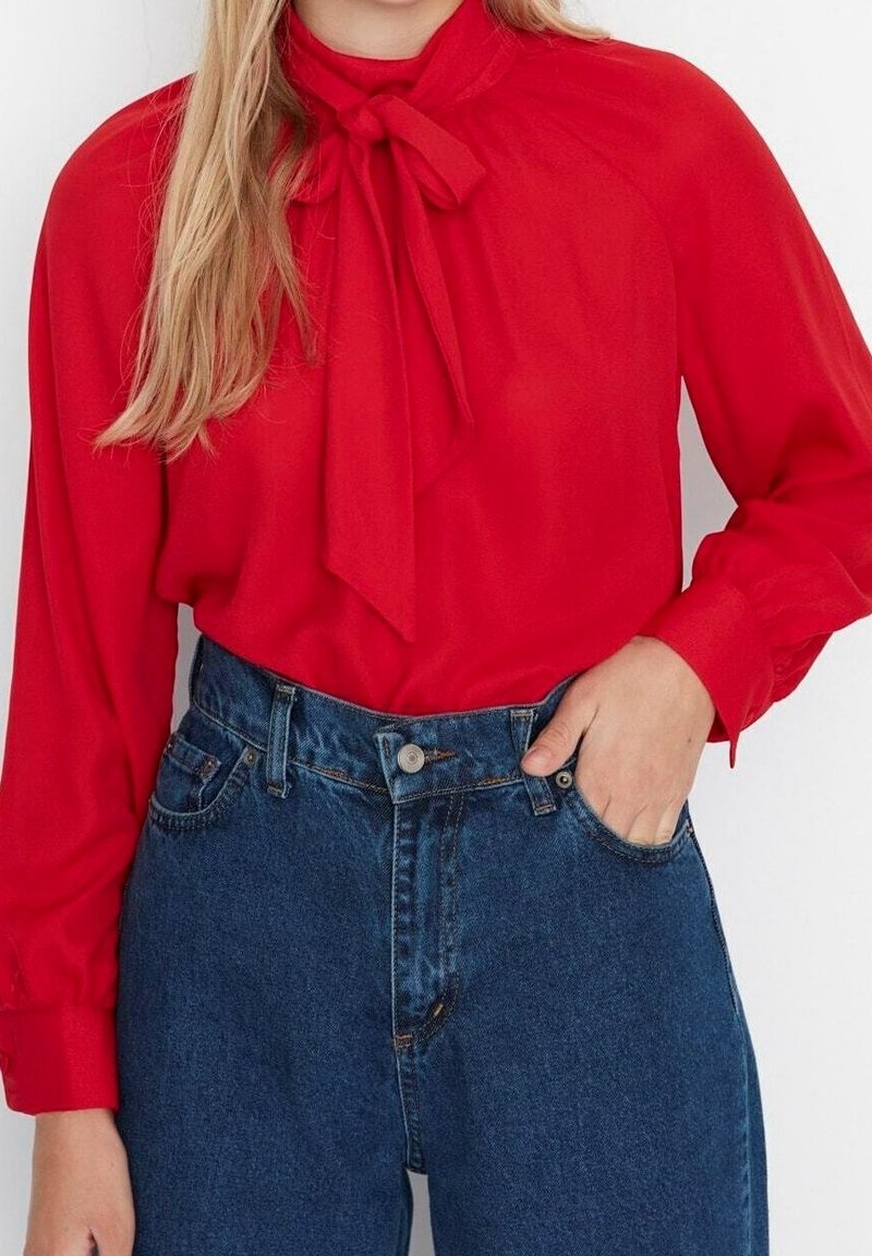 Femme portant un chemisier rouge vif à manches longues avec un nœud noué au col et un jean bleu taille haute, main dans la poche.
