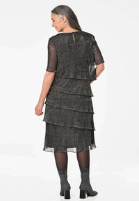 Schwarz geschichtetes Kleid aus texturiertem Stoff, mit drei Lagen, kurzen Ärmeln und einem Schlüsselkernausschnitt auf der Rückseite. Kombiniert mit grauen, strukturierten Socken.