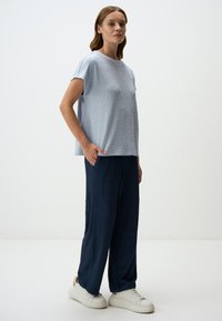 Lichtblauw gemêleerd topje met korte mouwen, gecombineerd met losse marineblauwe broek. Witte sneakers maken de outfit compleet, tegen een neutrale achtergrond.