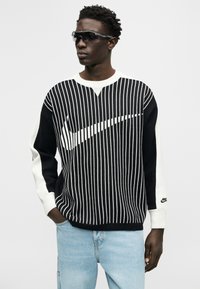 Νέος άνδρας που φοράει μαύρο και λευκό ριγέ πουλόβερ Nike με μεγάλο σήμα swoosh, ανοιχτόχρωμο τζιν και σκούρα γυαλιά ηλίου, κοιτάζοντας πλάγια.