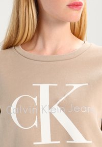 Felpa beige con scollatura ampia, caratterizzata da un evidente logo bianco "CK" e dalla scritta "Calvin Klein Jeans" in un carattere moderno. Texture liscia.