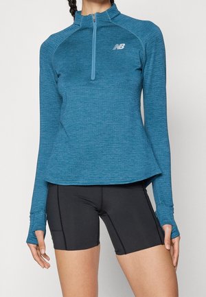 Kobieta ubrana w niebieską sportową bluzkę z długim rękawem z otworami na kciuki oraz czarne dopasowane szorty, widoczna od klatki piersiowej do połowy uda.