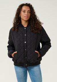Veste bomber noire matelassée avec un col et des poignets côtelés, dotée de boutons-pression et de poches latérales, associée à un jean bleu clair.
