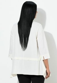 Persona con lunghi capelli neri lisci, indossa una maglietta bianca a maniche a tre quarti e pantaloni neri, vista di spalle su uno sfondo bianco uniforme.