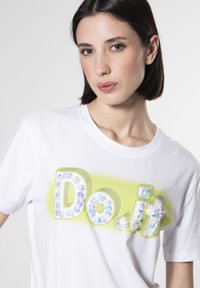Hvid t-shirt med teksten "Do.it" udsmykket med farverige perler og rhinsten, fremhævet af en lys gul gradient baggrund.