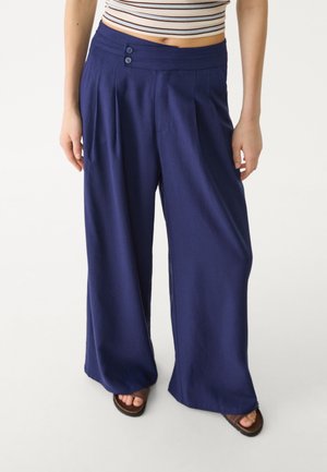 Kobieta ubrana w granatowe, szerokie spodnie z wysokim stanem z detalami guzików, zestawione z pasiastym topem odsłaniającym brzuch i sandałami.