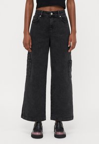 Calças cargo de perna larga em denim preto com bolsos laterais, fecho de botão e um desvanecimento subtil. Usadas com sapatos de couro pretos com um acabamento brilhante.