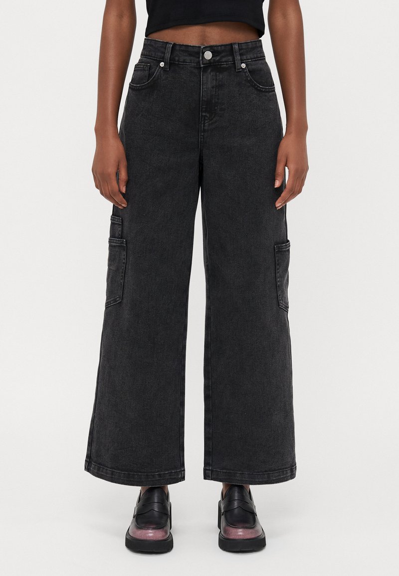 Calças cargo de perna larga em denim preto com bolsos laterais, fecho de botão e um desvanecimento subtil. Usadas com sapatos de couro pretos com um acabamento brilhante.