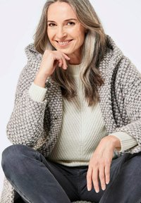 Cardigan surdimensionné en tricot gris et blanc avec une capuche, présentant un tissu texturé et des détails de boutons, associé à un pull côtelé crème clair.