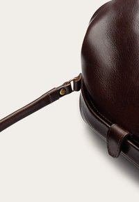 Ruda odinė rankinė su apvalia forma, pasižyminti lygiu paviršiumi, reguliuojama dirželiu ir auksiniu aksesuaru prie tvirtinimo taško.