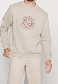 Beige mikina se žebrovanými manžetami a spodním lemem, s kulatým logem v hnědém písmu a vavřínem. Měkká, bavlněná smíšená tkanina.