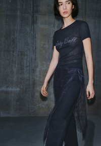 Crna, uska majica s kratkim rukavima s natpisom "party call", uparena s prozirnim tamnoplavim čipkastim omotačem oko tamnoplavih hlača. Ležeran, slojeviti izgled.