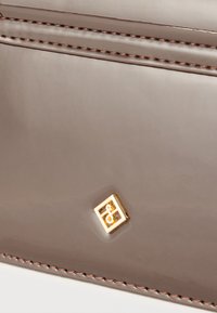 Γυαλιστερή επιφάνεια από δέρμα ταουπέ με καφέ ραφές και ένα μικρό χρυσό τετράγωνο έμβλημα με ένα στυλιζαρισμένο σχέδιο "S".