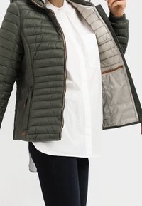 Dämpad, olivgrön jacka med ett kviltat mönster, med dragkedja fram, beige innerfoder och bruna hårdvaruaccenter.